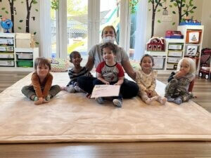 A small group of smiling children are sitting in a line on a carpet on the ground, alongside their care provider--flooring and carpeting