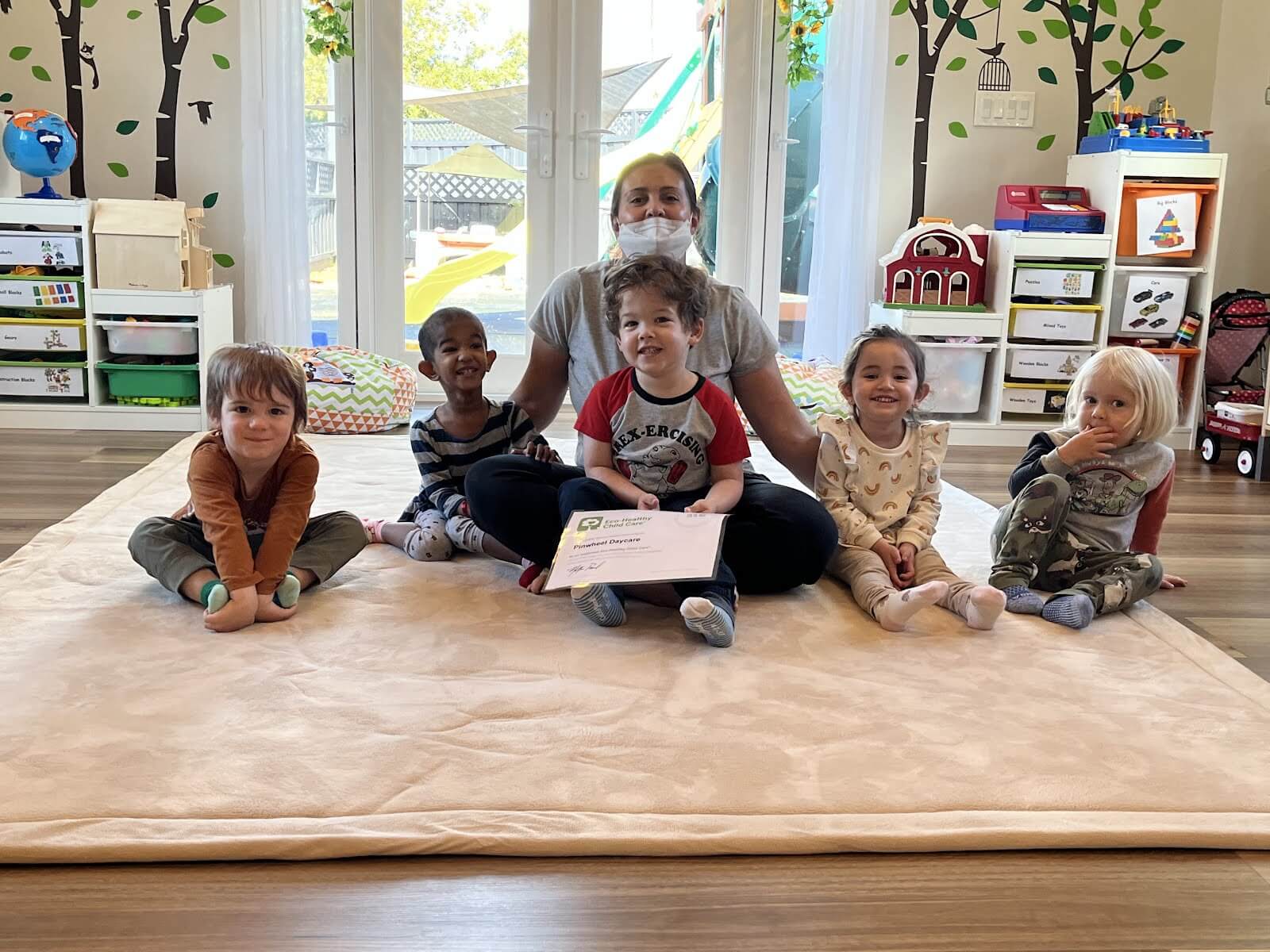 A small group of smiling children are sitting in a line on a carpet on the ground, alongside their care provider--flooring and carpeting