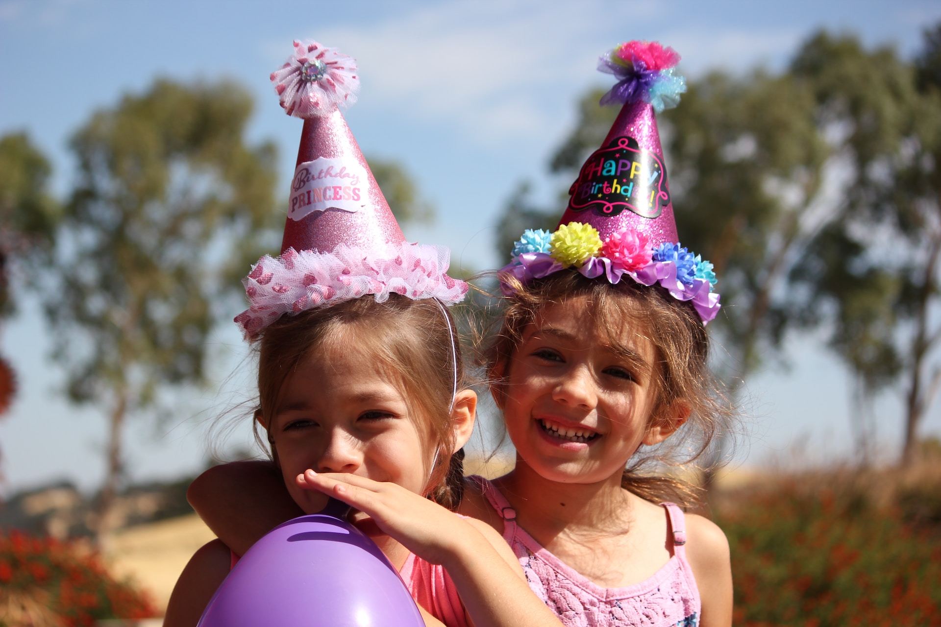 Two girls are standing outside with arms wrapped around the other's shoulder, smiling, wearing party hats, and holding a balloon--Celebrations