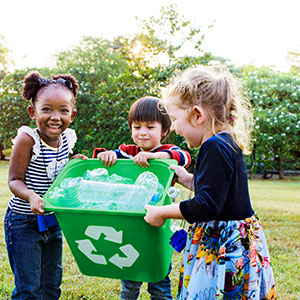 Three kids are outside holding a green recycling bin with plastic water bottles inside--Reduce