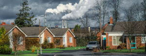 A picture with brick houses lined up in the foreground and industrial stacks in the background, streaming emissions into the cloudy sky