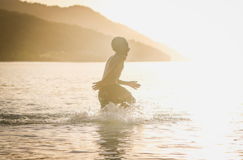 Boy runs through natural body of water in sunshine in front of mountains