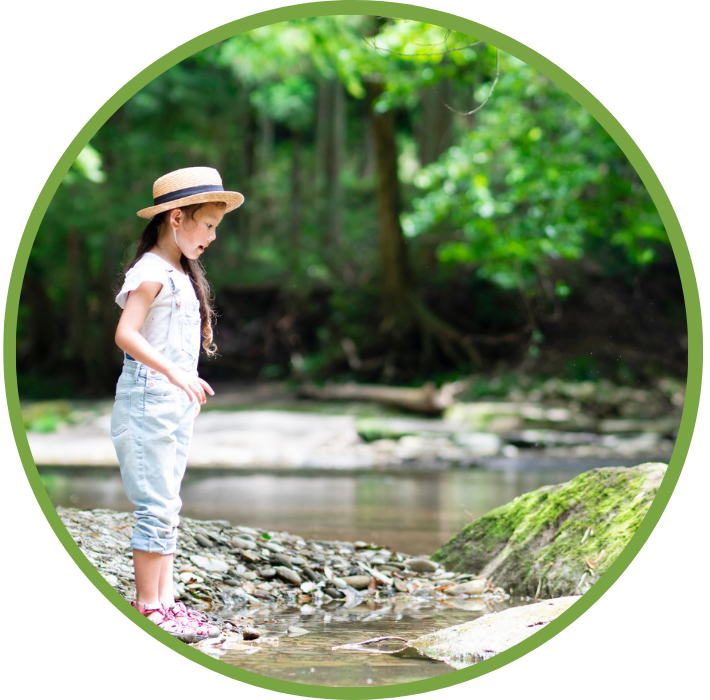 A young Asian girl stands on the side of a creek and looks at the water, surrounded by green trees