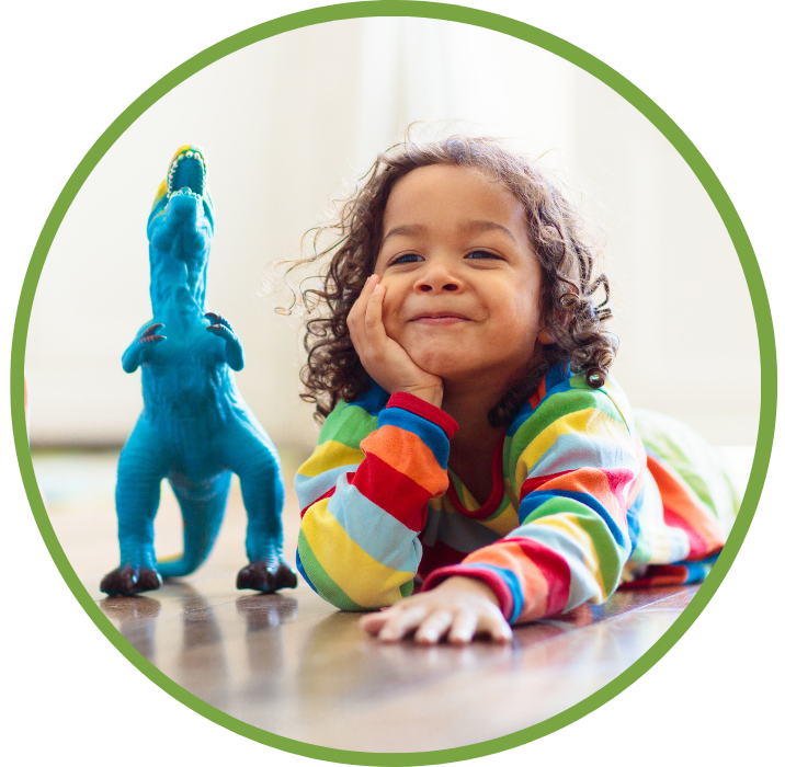 A young latina girl lays on the floor in a bright sweater, grinning, beside a bright blue toy T-Rex