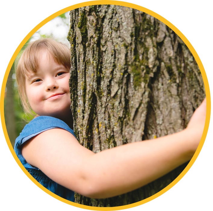 A young white girl with down syndrome hugs a tree and smiles