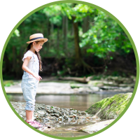 A young Asian girl stands on the side of a creek and looks at the water, surrounded by green trees