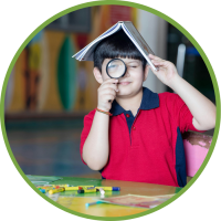 A young boy in a red shirt poses holding a magnifying glass in front of his face while balancing an open book on his head